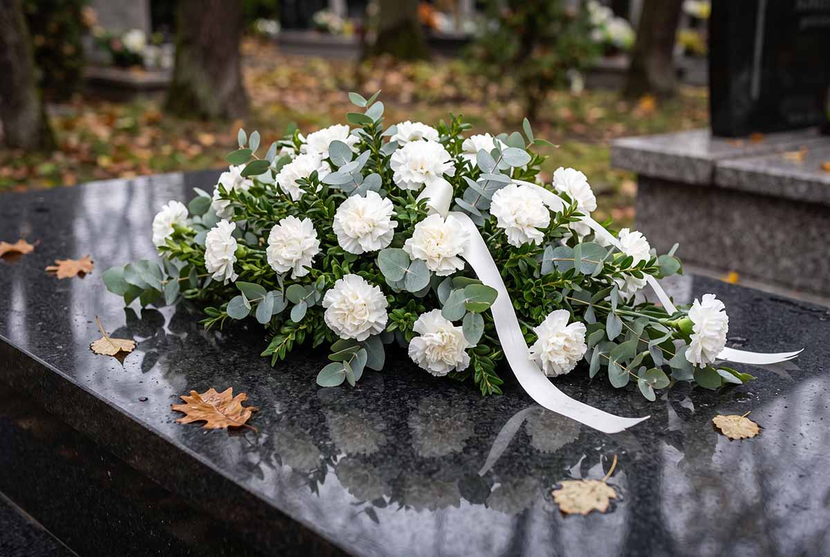 Elegancka kompozycja w bieli i zieleni na cmentarz Biało-zielona kompozycja na grób z goździków, eukaliptusa i bukszpanu na ciemnym granicie